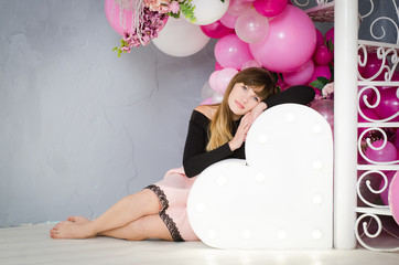 Beautiful girl sitting next to the heart with the light bulb. White wrought iron spiral staircase decorated with pink balloons and bouquets of roses