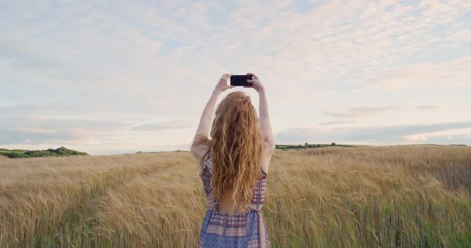 Rear view of attractive red head woman taking photograph wheat field using smartphone capturing epic nature background 