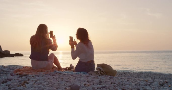 Two girl friends taking photograph of sunset on empty beach smartphone women photographing sunrise scenic landscape nature background view enjoying vacation travel adventure