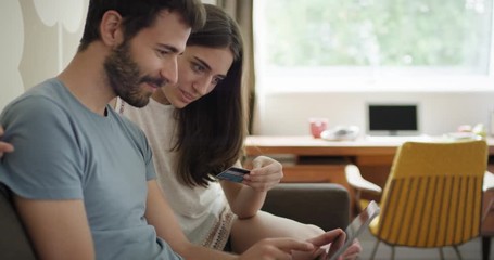 Young couple shopping online using credit card payment purchasing on digital touch screen together sitting on sofa celebrating with a kiss - Powered by Adobe