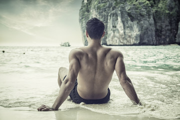 Full body back shot of a handsome young man sitting on a beach in Phuket Island, Thailand, shirtless wearing boxer shorts, showing muscular fit body