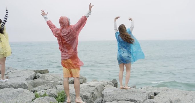 Diverse group of teenage friends arms raised wearing multi colored raincoat poncho in rainy weather on the beach celebrating vacation adventure 