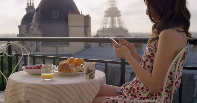 Beautiful young woman holding smartphone digital touchscreen sitting on Hotel Balcony enjoying sunrise view of Paris skyline Eiffel Tower in Background