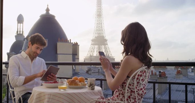 Young tourist couple in Paris hotel enjoying breakfast using smartphone tablets on terrace view of Eiffel Tower at Sunrise in the morning