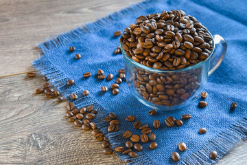 A backlit glass cup filled with roasted whole coffee beans on a blue woven placemat on a wooden...