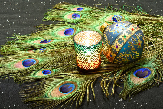 Exotic New Year or Mardi Gras image with peacock feathers, lit candle and ornate Christmas ball on a sparkly black fabric
