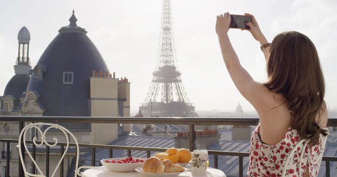 Woman taking photograph of Eiffel Tower using smartphone from hotel balcony at sunrise photographing scenic Paris cityscape background view enjoying European vacation travel adventure wearing pretty dress