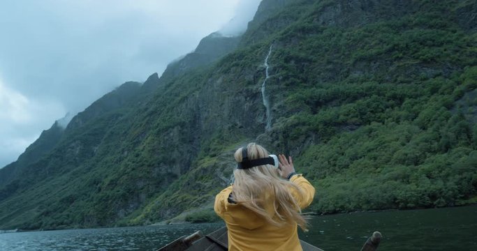 Brave woman traveler on row boat in Fjord Norway wearing virtual reality headset enjoying outdoor travel experience wearing yellow jacket  watching 360 video imagination concept