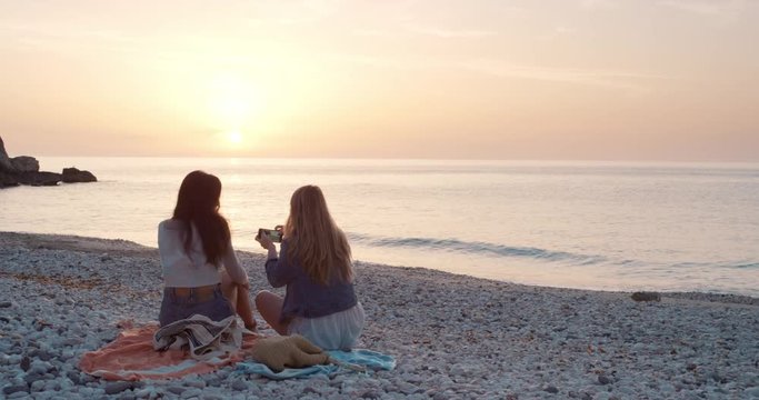 Two Women sitting on beach using smartphone smiling and texting best girl friends with mobile phone browsing social media for inspirational travel photos enjoying a relaxing summer vacation at sunset on beach