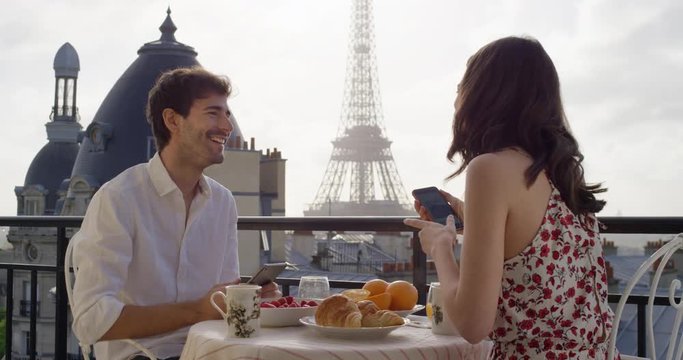 Young tourist couple in Paris hotel enjoying breakfast on terrace woman showing holiday photo to happy  man view of Eiffel Tower at Sunrise in the morning