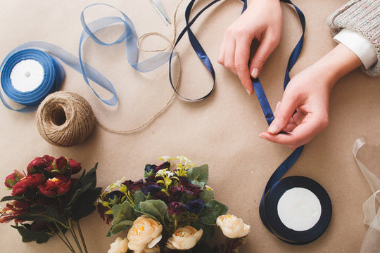 Preparing decorations for holiday, fancywork, handmade art. Unrecognizable woman hold blue ribbon on beige background with artificial flowers, tape rolls and rope, flat lay.