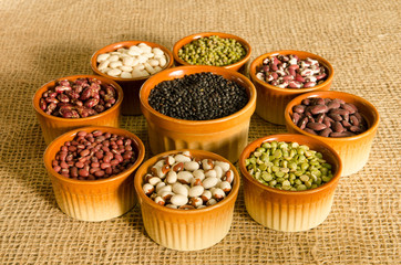 9 pottery containers with various types of pulse and legume seeds on a burlap background in late afternoon light.