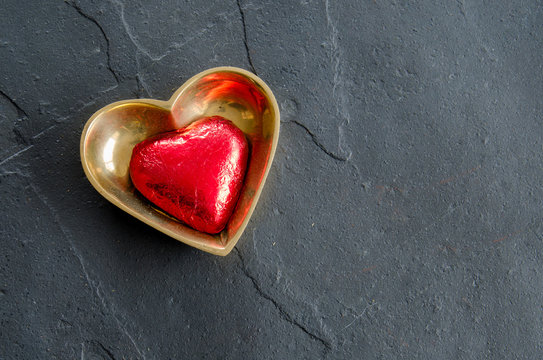 A red wrapped chocolate heart in a brass heart shaped dish on a black slate background for Valentine's Day