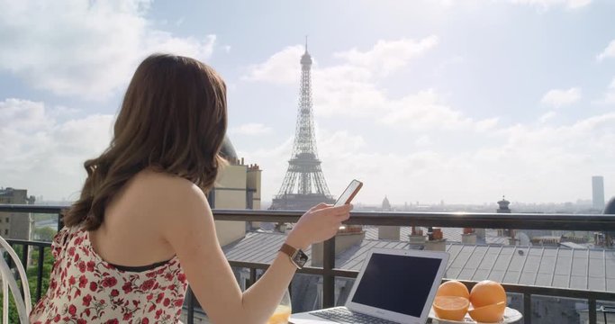 Beautiful young woman holding smartphone digital touchscreen sitting on Hotel Balcony enjoying sunrise view of Paris skyline Eiffel Tower in Background