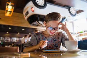 Beautiful attractive girl with glasses looks at her tablet computer. Female work in the interior of a stylish cafe. People and the Internet.