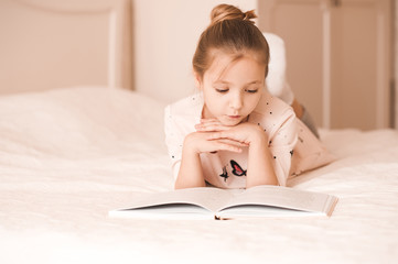 Cute baby girl lying in bed reading book in room. Wearing pajamas at home. Childhood.