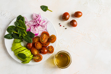 White plate with ingredients for healthy vegetarian salad, sliced cherry tomatoes, red onion, avocado and spinach leave on the concrete background, top view.