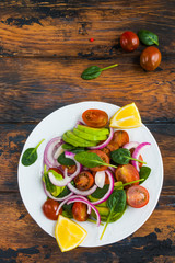 Vegetarian salad with fresh cherry tomatoes, red onion, avocado and spinach leaves on a white plate on the wooden rustic table, top view.