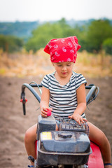 child in red bandana and stripe tee  sits on tiller on the field. baby girl driver cultivator