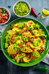 Nachos with cheese, jalapeno peppers, red onion, parsley, tomato, salsa, guacamole sauce and tequila on green plate.