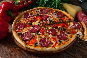 Appetizing sliced italian pizza with salami sausage, tomatoes, parmesan cheese, pepper, parsley and olives served on wooden background. Restaurant menu photo.