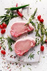 Raw Pork Loin chops on a cutting board with herbs, rosemary, thyme, chilli, salt, pepper on white cutting board.