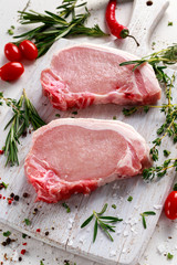 Raw Pork Loin chops on a cutting board with herbs, rosemary, thyme, chilli, salt, pepper on white cutting board.