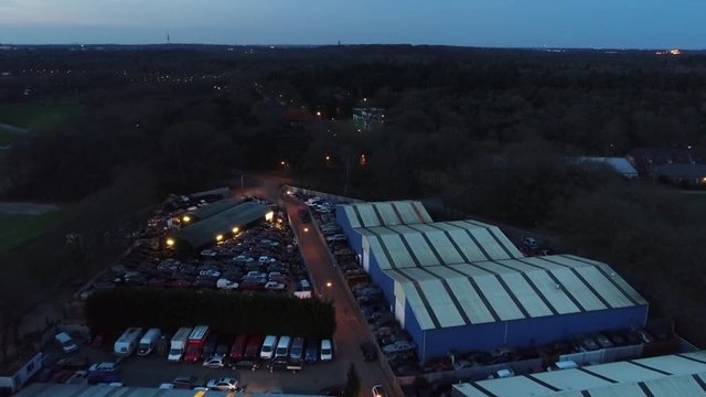 Aerial At Night Flying Over Scrapyard On Left Showing Piled Up Scrap Cars And On Right Wrecking Yard Warehouses Where Recycling Is Done Dark Evening Some Lights Seen On Horizon And Background 4k