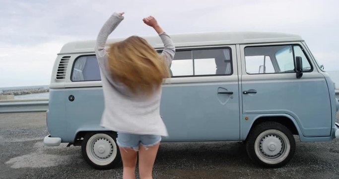 Happy hipster woman dancing freestyle dance at festival in front of retro camper van celebrating travel adventure