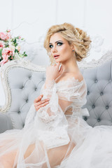 Fototapeta premium Beautiful blonde girl sitting on a sofa dressed in a white negligee holding a wedding bouquet, sending an air kiss in the background many colors