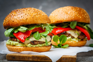 Tasty grilled beef burger with cheese, guacamole and pickled peppers. Dark background, brown paper, wooden stand.