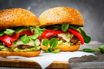 Tasty grilled beef burger with cheese, guacamole and pickled peppers. Dark background, brown paper, wooden stand.