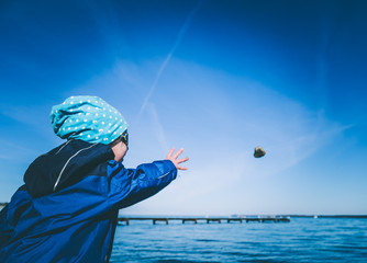 kleines Kind schmeisst Stein am Strand vor blauem Wasser weit durch die Luft