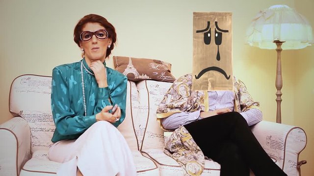 A logorrheic harsh wife keeps talking and watching the tv, while the husband saddens (he wears a breadbag face). Funny comedic shot.
