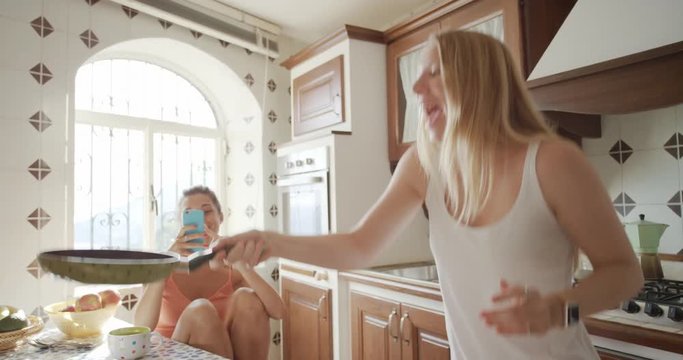 Best Friends making home made Pancakes girl taking photo using smart phone Lifestyle Woman flipping flapjack at home sharing on social media