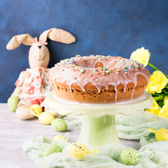 Easter bundt ring cake with sugar frosting sprinkles decorations eggs and doll bunny on dark blue background. Festive holiday home made treat