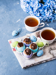 Easter sweet cup cakes treets in colorful pastel egg shells with chocolate and hydrangea flowers on blue background. Festive holiday home made food with cups of tea