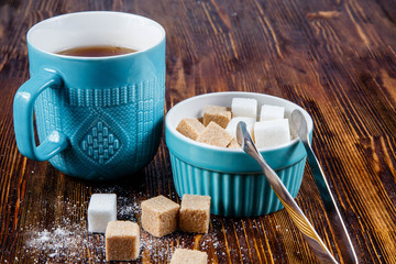Close-up of a blue ceramic mug with tea and a sugar bowl with cane and white sugar on a wooden table.