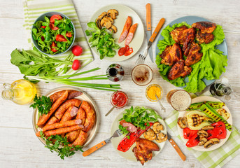 Dinner table with variety food, fried chicken wings, sausages, grilled vegetables in a pan, salad and  lager beer on wooden table, top view. Indoor food Concept.