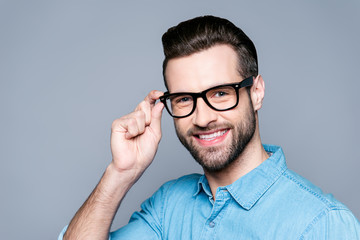 Portrait of young smiling manager in jeans shirt touching his glasses isolated on gray background