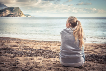 Pensive lonely young woman sitting on beach hugging her knees and looking into the distance with...