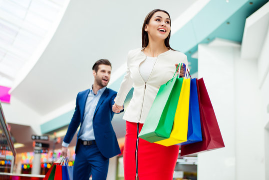 Hurry faster! It's shopping time and fun. Handsome cheerful  successful happy  man  holding hands and following his lovely cute woman with colored shopping packages and laughing in mall at holiday.