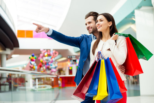 It's shopping and fun  time. Cheerful  successful happy young lovely couple in jacket holding  colored shopping bags, pointing at shop and laughing in mall at holiday