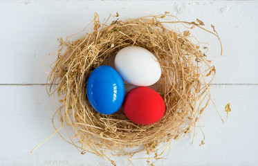 Fototapeta premium Red, blue, white eggs ( As flag of some countries ) in nest on rustic wooden background, selective focus image. Happy Easter Card - space for text. 