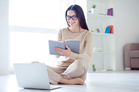 Portrait of young cheerful woman in glasses sitting on the floor at home using laptop for styding online and writing information in her notepad