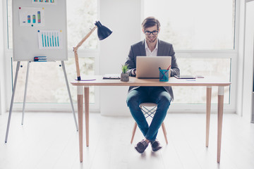 Portrait young handsome businessman with glasses is sitting at desk in workstation, smiling and typing on laptop