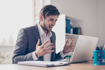 Angry young bearded businessman is sitting at a desk in the office, feeling nervous and making angry at his workstation