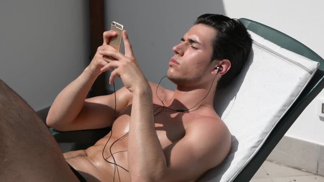 Shirtless muscular handsome young man sunbathing on sunchair, while listening to music through headphones