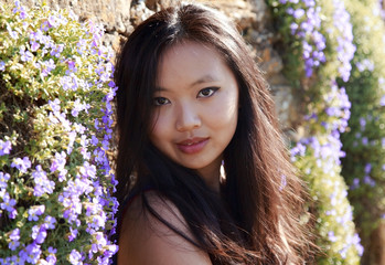 Naklejka premium Beautiful potrait of a Chinese, Oriental young woman with long dark hair sitting against a wall of purple flowers