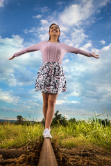 Fototapeta premium Cheerful young woman with outstretched arms is walking on railroad track
